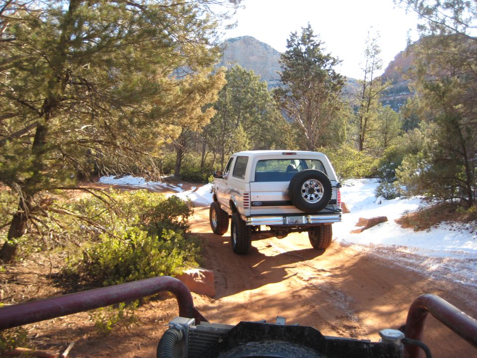 White 1996 Ford Bronco with spare tire on rear door driving on a dirt road with snow patches