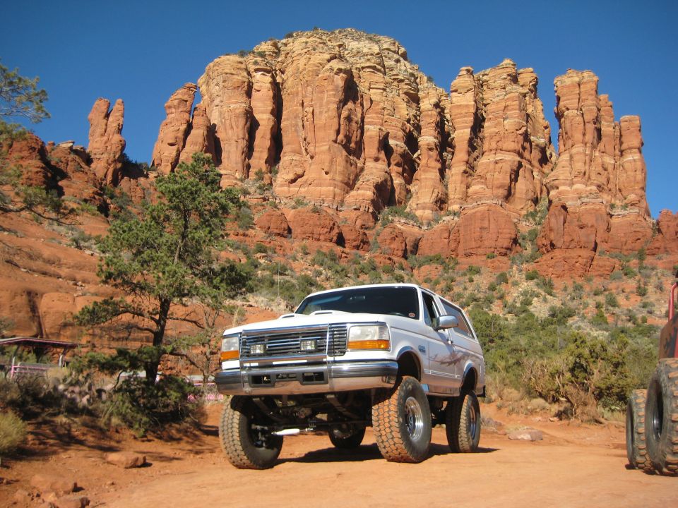 White Ford Bronco with lifted suspension and large off-road tires parked in desert landscape. Another vehicle is partially visible on the right