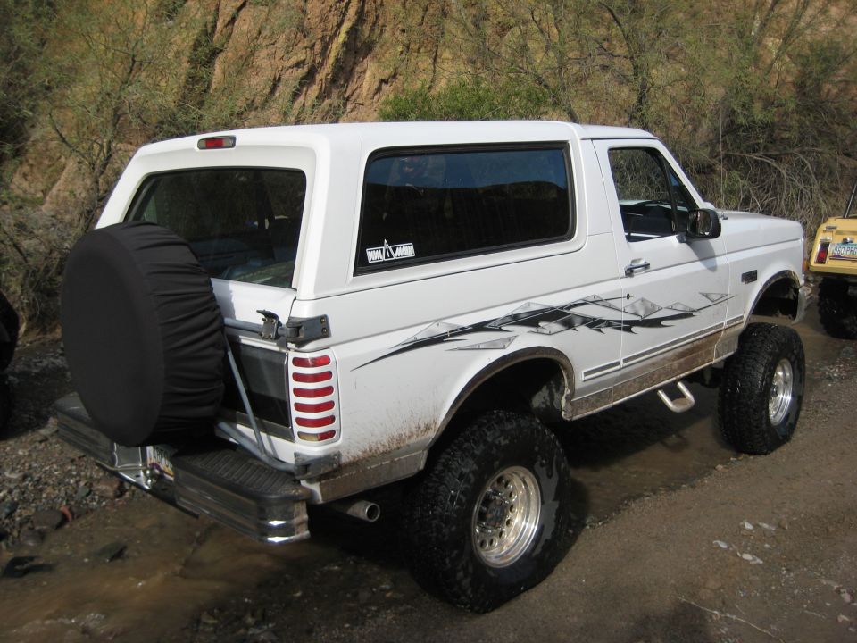White Ford Bronco with aftermarket graphics and large off-road tires parked on dirt. Another yellow vehicle is partially visible to the right
