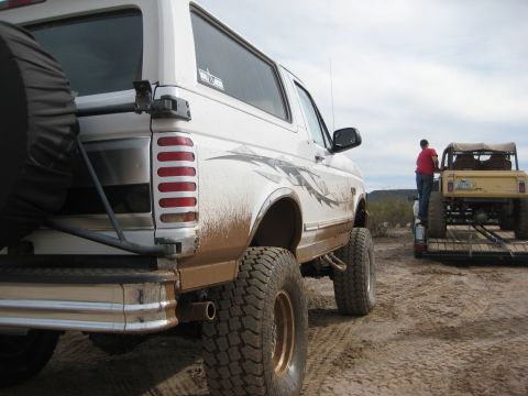 1996 Ford Bronco - A white Ford Bronco, caked in mud, is parked next to a trailer carrying a yellow Jeep in a dusty, outdoor setting.