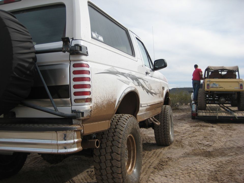 White Ford Bronco, rear quarter view, covered in mud. Another vehicle on a trailer behind it
