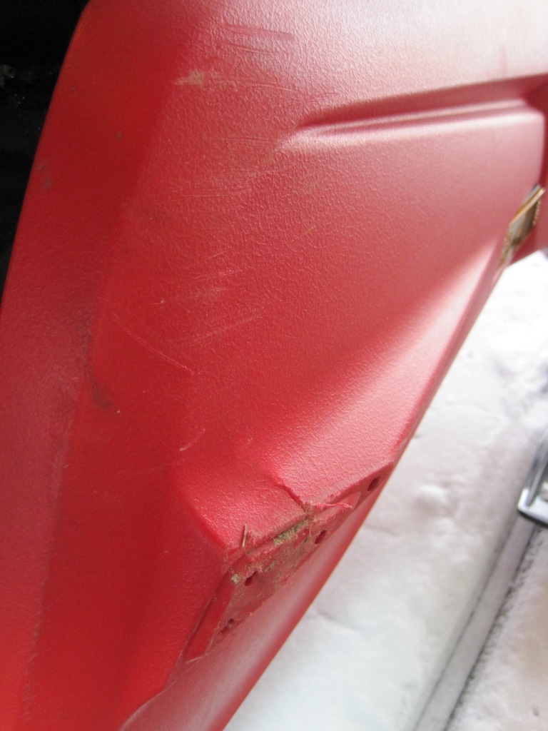 Cracked and scuffed red plastic snowmobile side panel close-up, with snow in the background