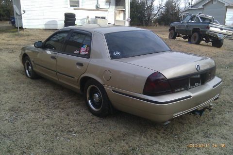 2000 Mercury Grand Marquis - A tan sedan is parked on a grassy area next to a house, with a lifted pickup truck visible in the background.