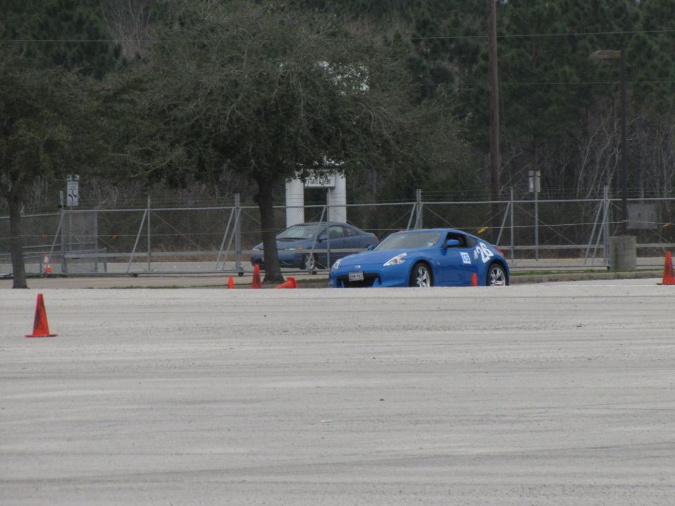 Blue sports car with number 26 on door, parked on a dirt lot with orange cones