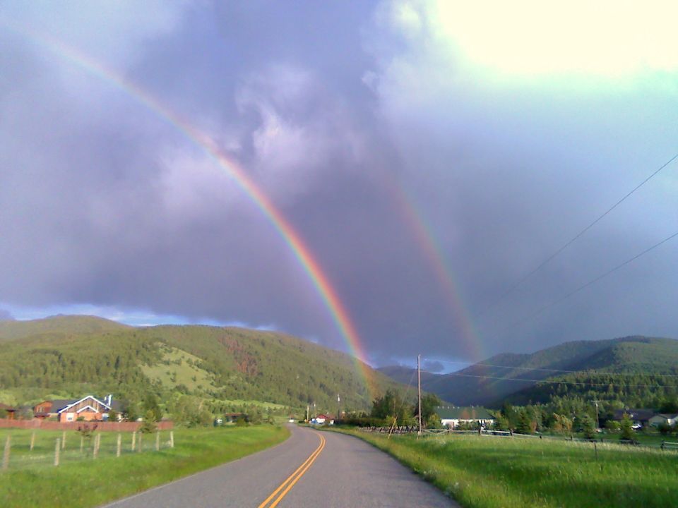 Double rainbow arcs across a cloudy sky over a two-lane road in a green, hilly landscape with houses visible
