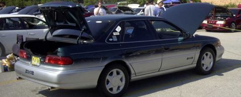 1997 Ford Taurus - A dark green and silver Ford Taurus is shown with its trunk and hood open, parked in an outdoor lot with other vehicles.
