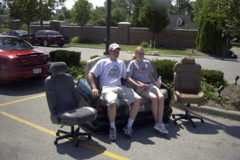 1997 Ford Taurus - A man and a woman sit on car seats placed in a parking lot, with a red car visible behind them.