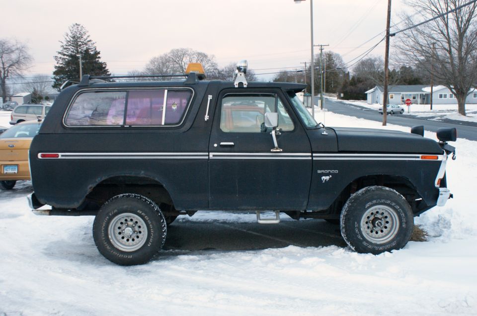 Black 1978 Ford Bronco parked in snow, side view. It has a roof rack and a yellow beacon light