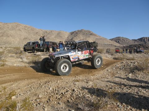 1986 Ford Bronco - A custom off-road buggy navigates a dusty desert course, with other vehicles and spectators in the background.