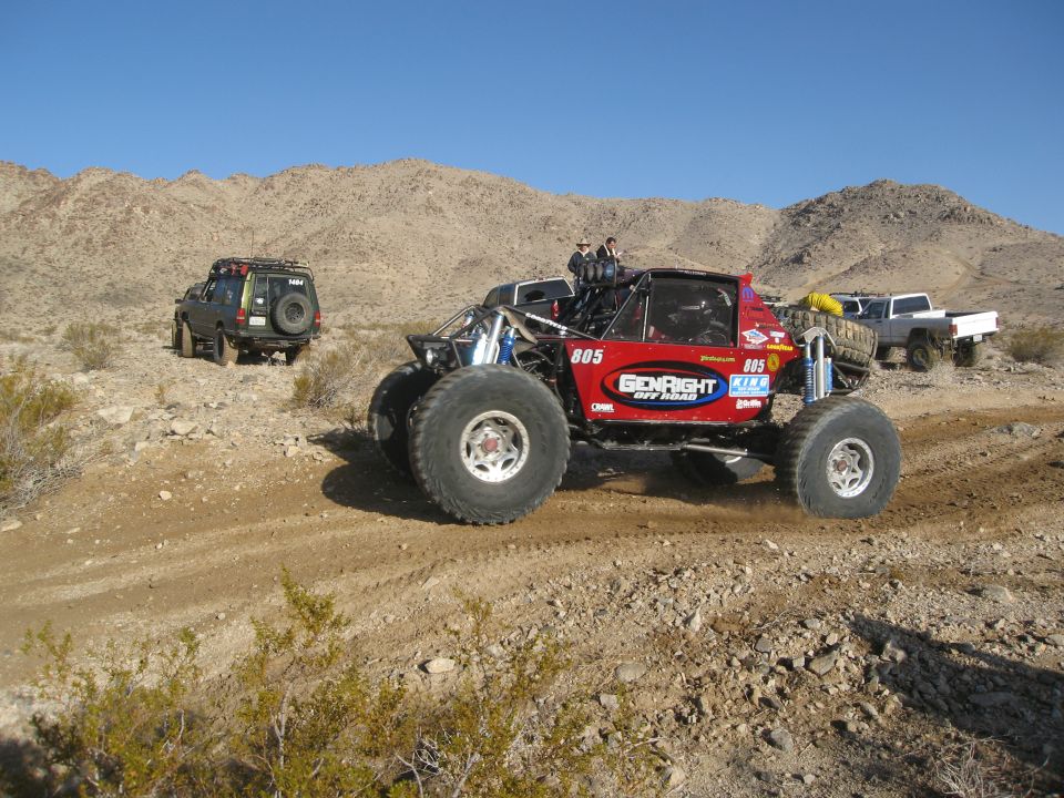 Red off-road buggy with large tires and "GENRIGHT OFF ROAD" livery, driving on a dirt track with other vehicles and desert landscape