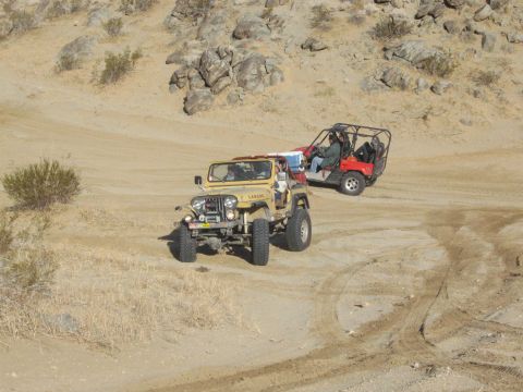 1986 Ford Bronco - Two off-road vehicles navigate a sandy desert landscape with rocky hills in the background.