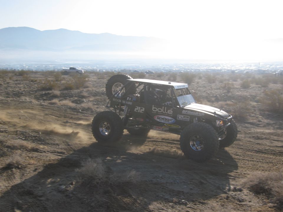 Black off-road buggy with large tires kicking up dust on a dirt track, spare tire mounted on the back