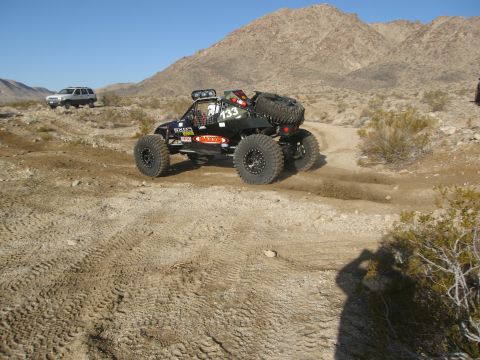 1986 Ford Bronco - A custom off-road buggy navigates a dusty desert trail, with another SUV visible in the background.