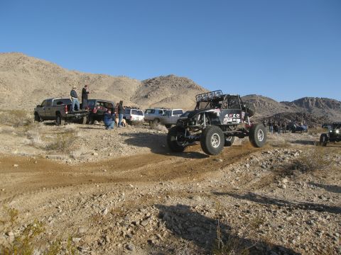 1986 Ford Bronco - An off-road buggy navigates a dusty trail in a desert landscape with several other vehicles and spectators present.