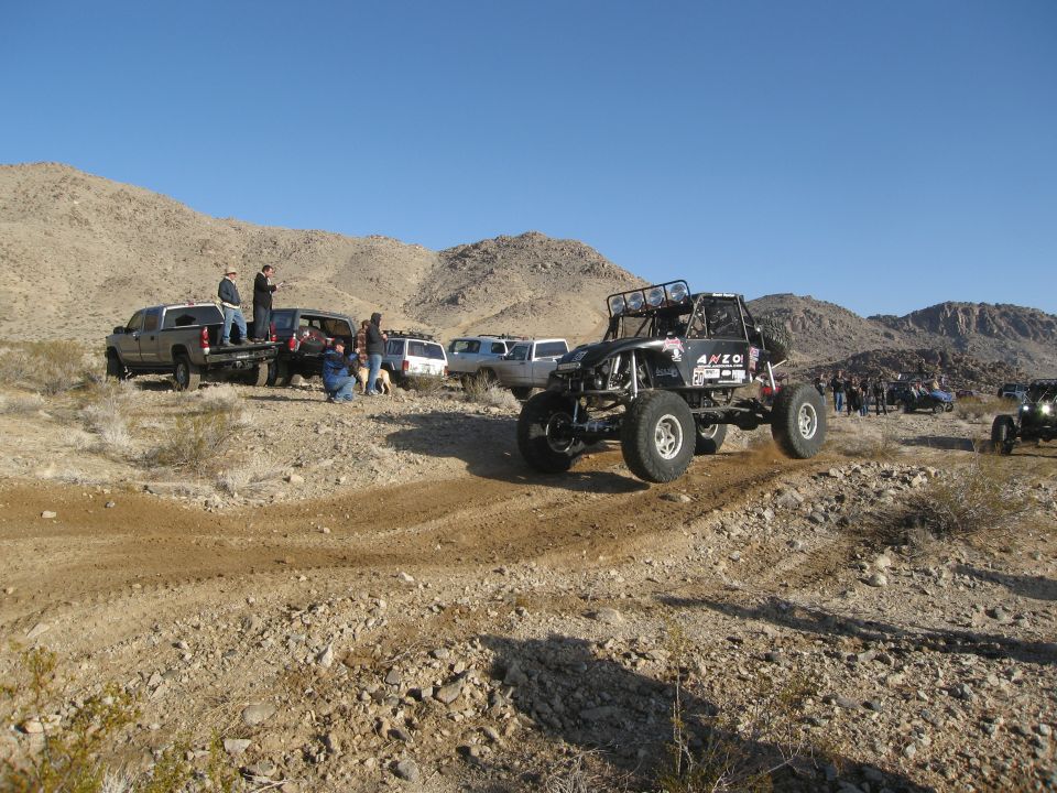 Buggy kicking up dirt on a desert trail with people and other vehicles watching