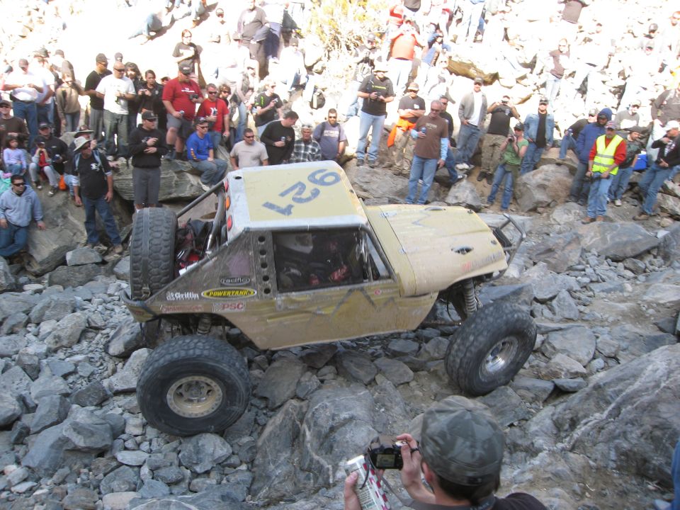 Off-road buggy with number 927 on top, navigating a rocky trail with a crowd watching