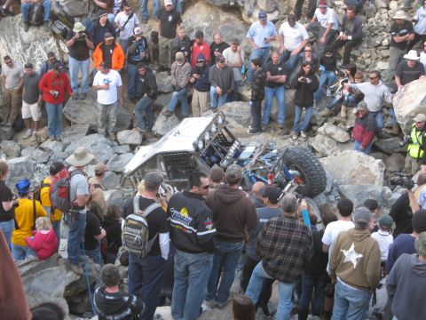 1986 Ford Bronco - A crowd watches an off-road buggy that has tipped onto its side amidst a rocky terrain.