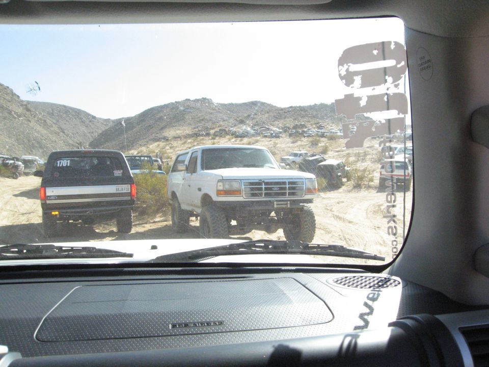 White Ford Bronco with lifted suspension and off-road tires, parked on a dirt road with other vehicles in the background
