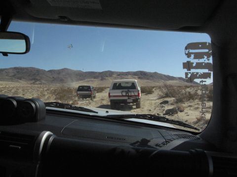 1986 Ford Bronco - Two SUVs traverse a dusty desert trail under a clear blue sky.