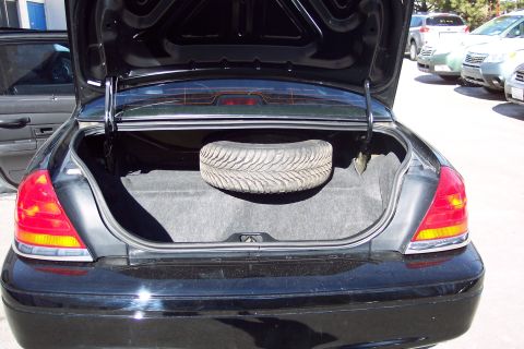 2001 Ford Crown Victoria - The open trunk of a black sedan reveals a spare tire, with other vehicles visible in the background.