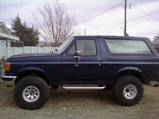1987 Ford Bronco - A dark blue 1987 Ford Bronco is shown from its side profile, parked outdoors with a fence and trees in the background.