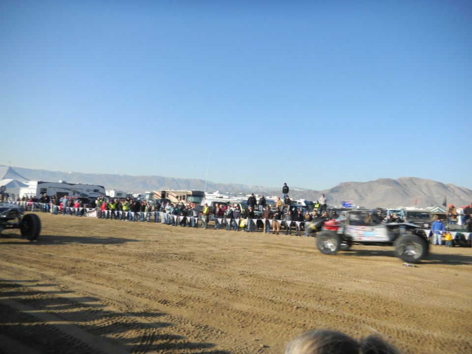 Off-road buggy races past spectators lined up behind a fence. Another buggy is visible on the left