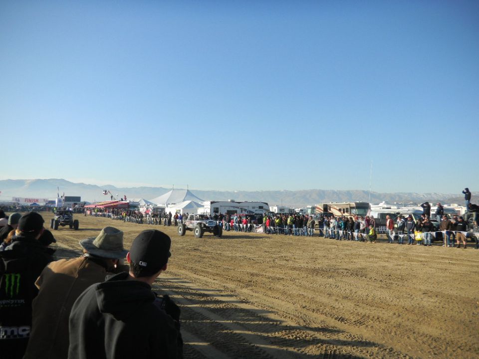 Off-road vehicles race past spectators in a sandy area under a clear blue sky