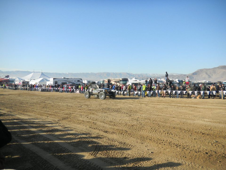 Off-road buggy with large tires drives on sandy terrain near spectators and tents