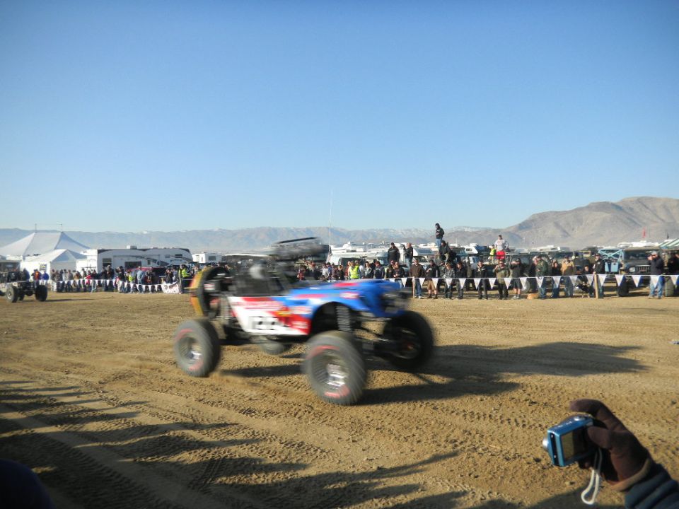 Off-road buggy with blue and red graphics, blurred by motion, on a sandy track with spectators