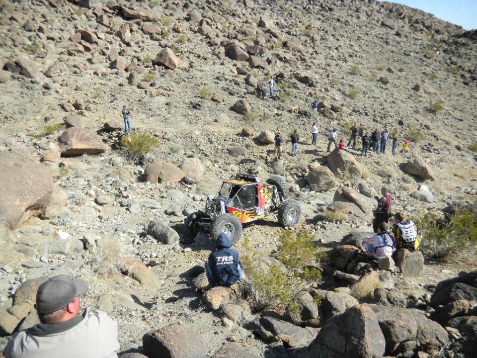 Off-road buggy with flame decals climbing a rocky hill with spectators watching