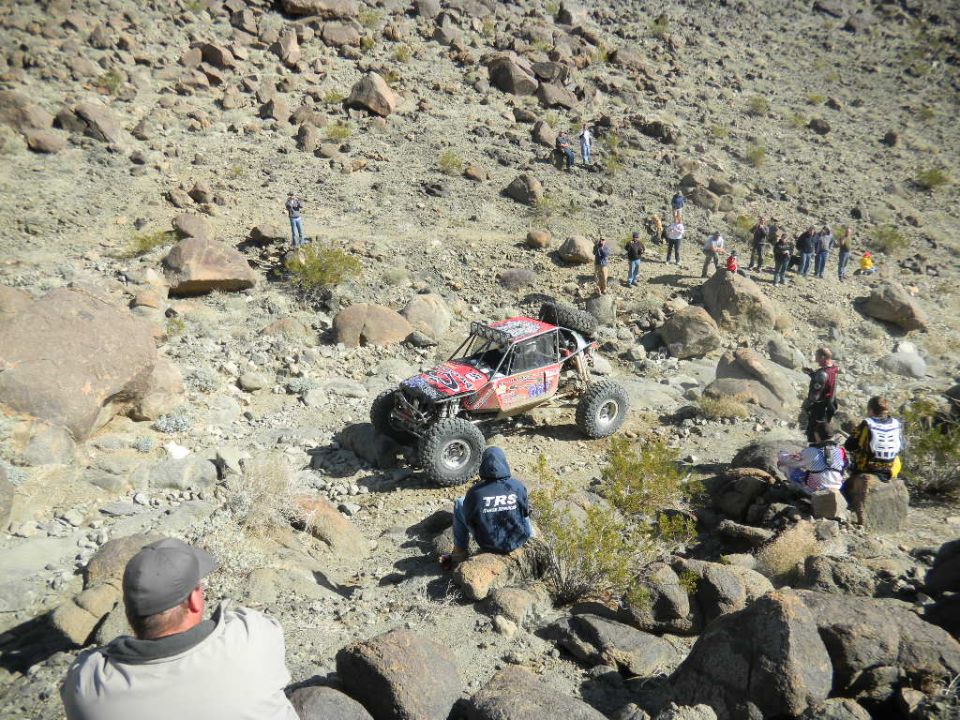Red off-road buggy navigates rocky terrain with spectators watching