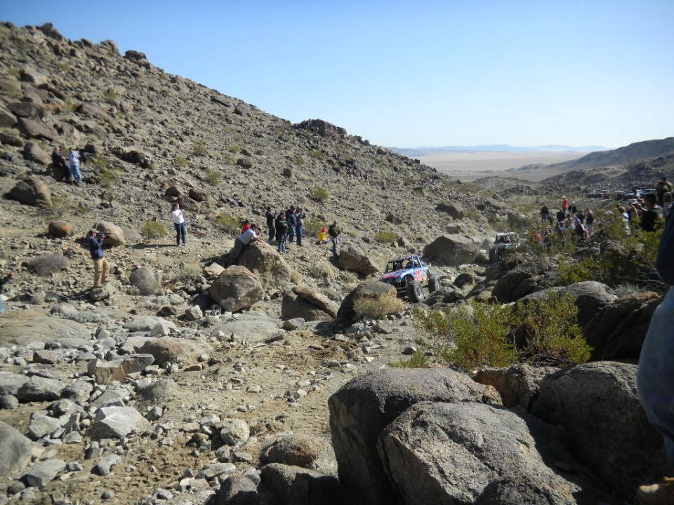 Off-road vehicles navigate a rocky desert terrain with spectators watching from hillsides