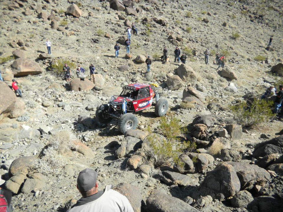 Red off-road buggy with large tires climbing a rocky hill. Spectators watch from the slopes
