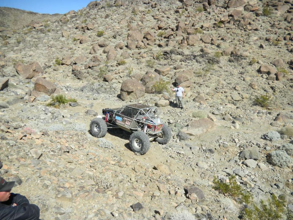 Off-road buggy with roll cage and large tires on a rocky desert slope. A person stands nearby