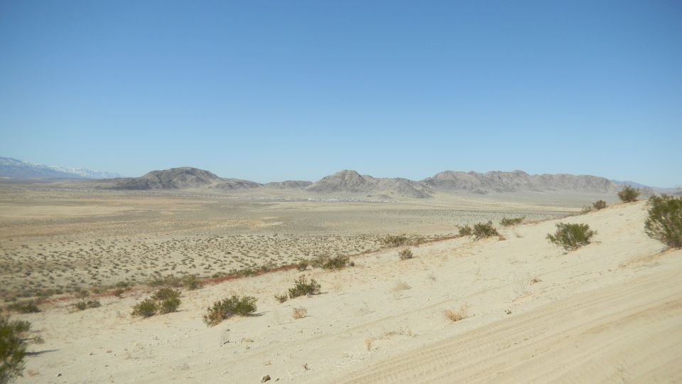 Desert landscape with scrub brush, sand dunes, and distant mountains under a clear blue sky. Supermotors.net logo in lower right
