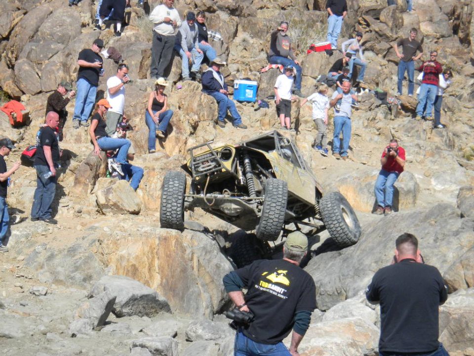 Off-road buggy with large tires climbing a rocky incline, spectators watch