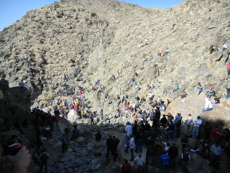 Spectators gathered on a steep, rocky mountainside under a clear blue sky