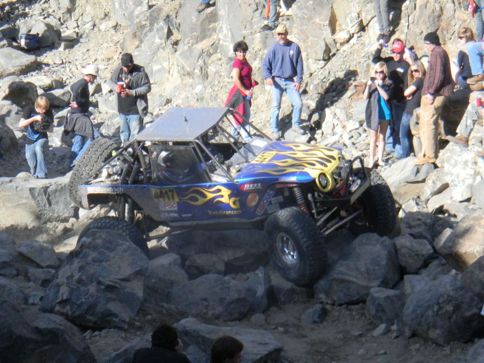 Off-road buggy with flames on hood navigating rocky terrain with spectators watching