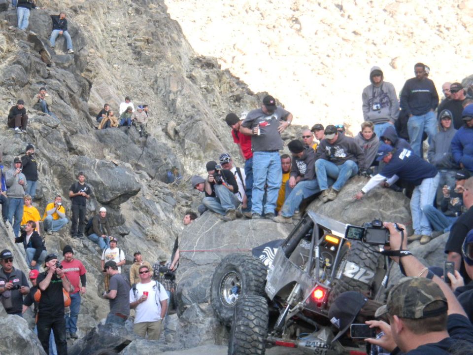 Off-road buggy with number 212 on side climbing a rocky incline, surrounded by spectators