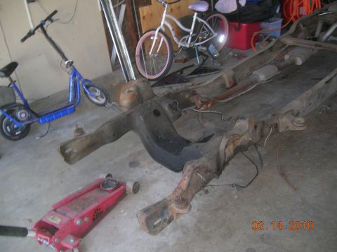 1985 Ford Bronco - The frame of a vehicle is visible on the ground, alongside a red hydraulic jack and a blue electric scooter.