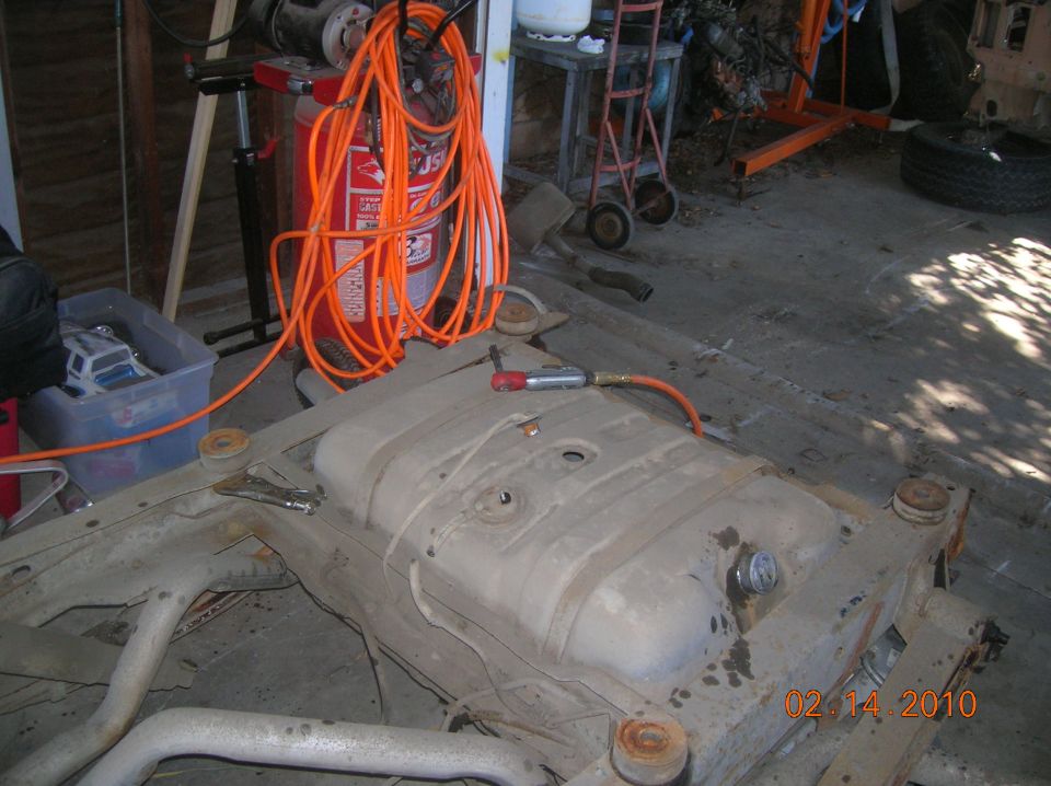 Vehicle fuel tank mounted on frame with exhaust pipes visible; air compressor and hose in background