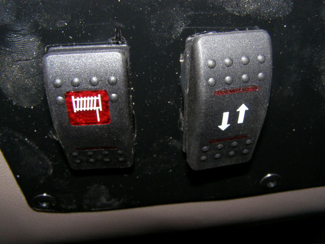 Two aftermarket rocker switches mounted on a dark dashboard. One has a red icon resembling a fence. The other has up and down arrows