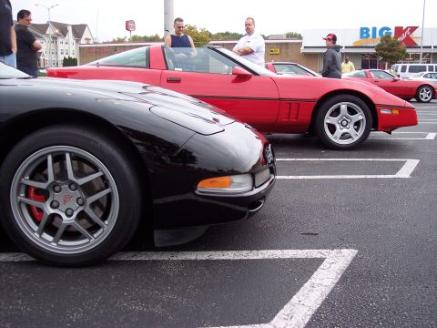 2006 Chevrolet Corvette - Two Chevrolet Corvettes parked in a lot with people nearby. The red Corvette appears to be a C4 generation model.