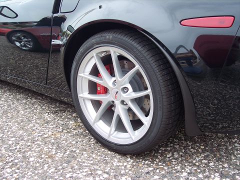 2006 Chevrolet Corvette - A black Chevrolet Corvette's rear wheel and side panel are shown, with a reflection of another red vehicle in the glossy paint.