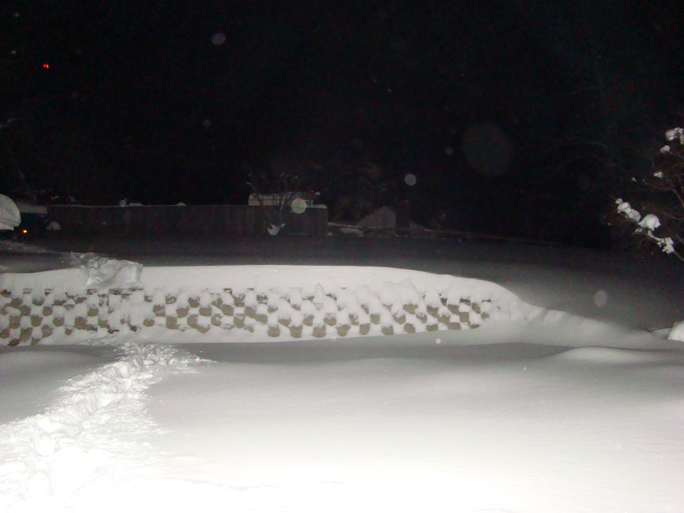 Snow covers a yard and a low wall at night. A watermark reads SUPERMOTORS.NET