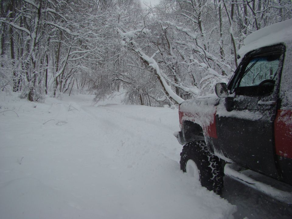 Red and black truck partially covered in snow, parked on a snow-covered road in a wooded area