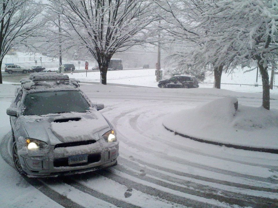 Snow-covered silver Subaru WRX wagon with roof rack and lights on in a snowy lot