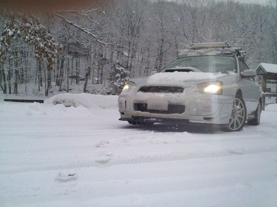 Snow-covered white Subaru Impreza sedan with roof rack and gold brake calipers in a winter driveway