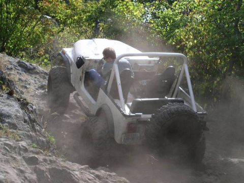 1979 Ford Bronco - White off-road vehicle navigates a dusty, rocky incline with trees in the background.