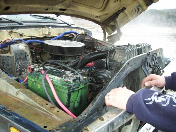 Engine bay with round air cleaner, green battery, blue hoses; person's arm resting on radiator support